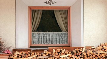 Cafe curtains inside the window above a wood pile.