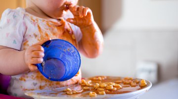 Mother Feeding Baby Sitting In High Chair At Mealtime