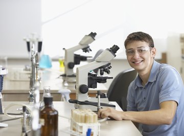 A young man in a science class sitting behind a microscope wearing glasses.