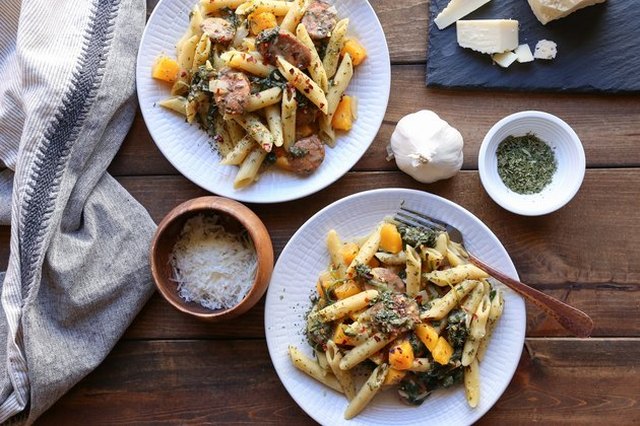 Overhead shot of two white plates of penne with squash and sausage on a rustic table, surrounded by the dish's ingredients and a clean kitchen towel
