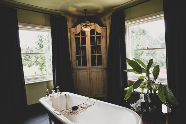 Interior view of bathroom with wooden corner cabinet between sash windows, roll top bath with brass bath caddy.