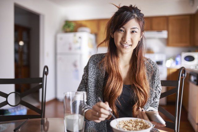 asian teen girl with smartphone in kitchen eating breakfast cereal