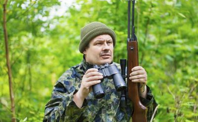A hunter holds binoculars and a rifle.