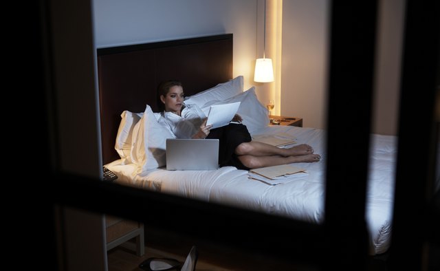 Businesswoman lying on bed reading papers, view through window, night