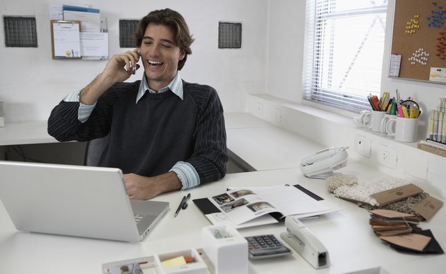 Young man talking on cell phone and using laptop at desk, laughing
