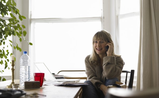 Young woman sitting at laptop on dining table, using cell phone, smiling