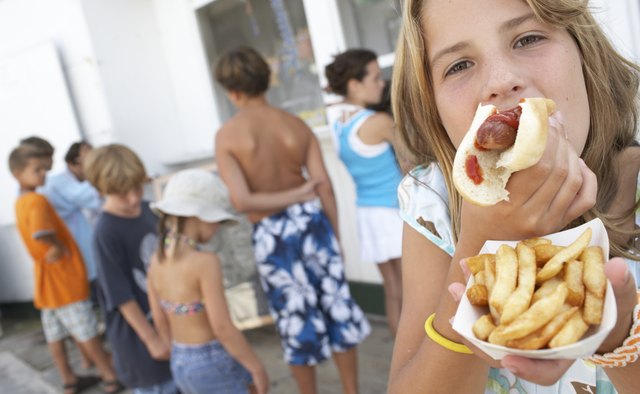 Girl (8-10) eating hotdog, children (6-12) queuing at vendor's window