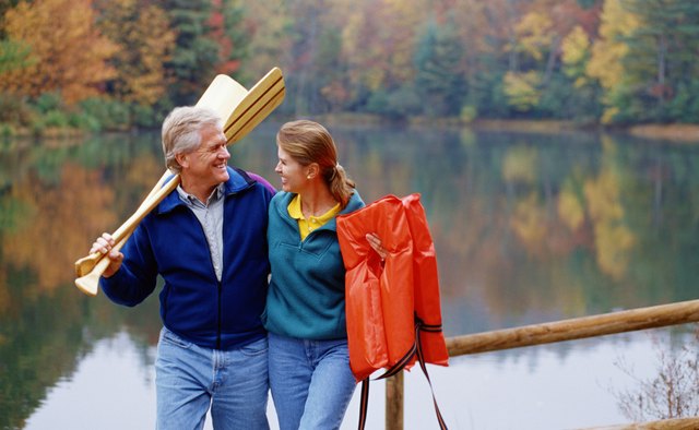 Couple walking by lake, man holding canoe paddles over his shoulder, woman holding lifejackets