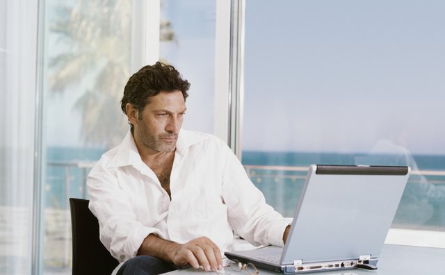 Mature Businessman Sits at a Table Indoors by a Patio Using His Laptop