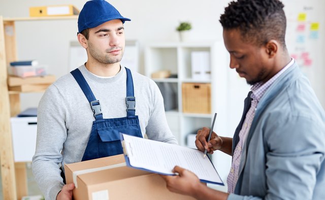 Serious calm young delivery man in cap holding cardboard box while black office manager signing for delivery in document