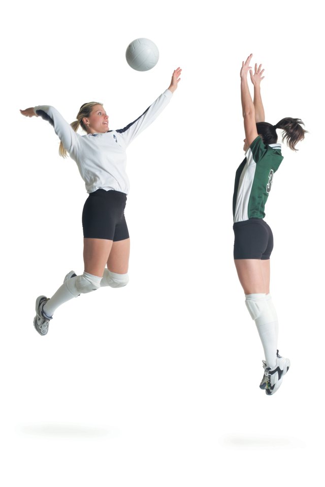 two young caucasian female volleyball players from opposing teams square off as one jumps and hits the ball while the other prepares to block