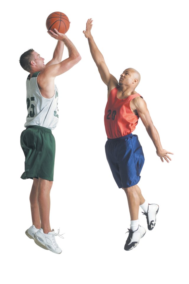 two young men dressed in opposing team basketball uniforms are jumping up while one prepares to shoot a basketball and the other tries to block it