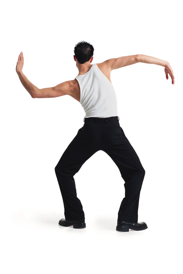 a young caucasian male dancer in black pants and white tank top stretches out and moves his arms with his back to the camera