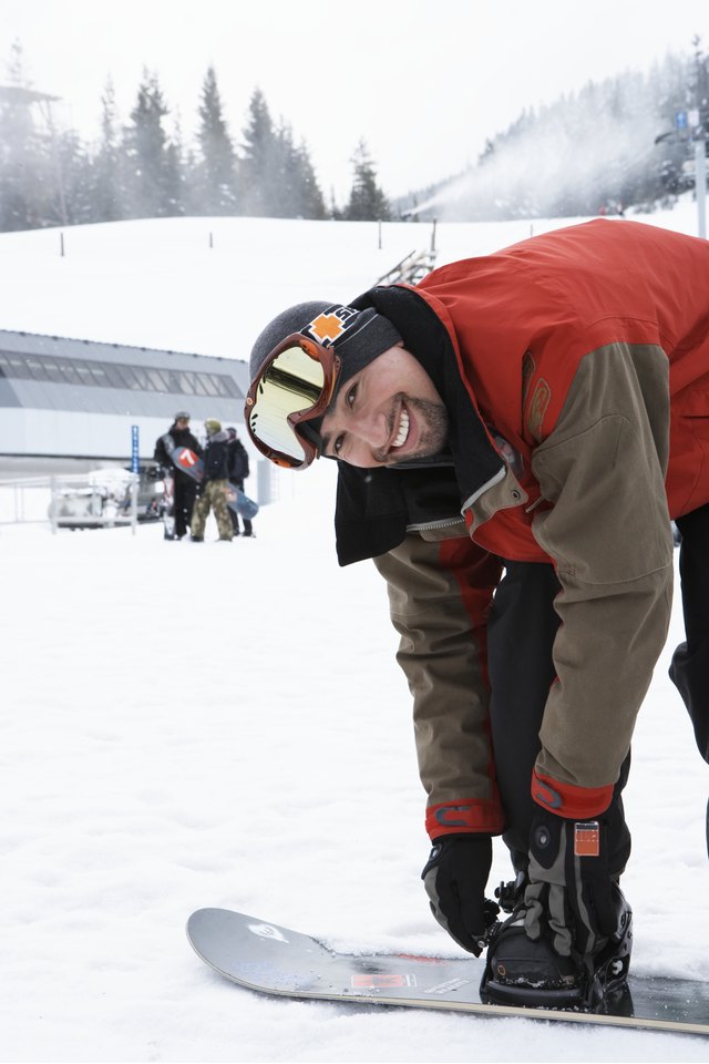 Man adjusting snowboard at base of mountain, smiling, portrait