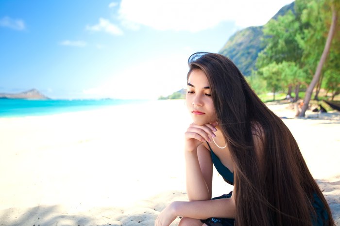 Young woman or teen girl sitting alone on beautiful Hawaiian beach looking sad or lonely, thinking, chin on hand