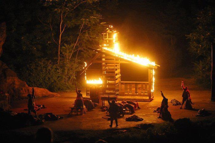 Actors surround a burning wooden house on an outdoor stage.