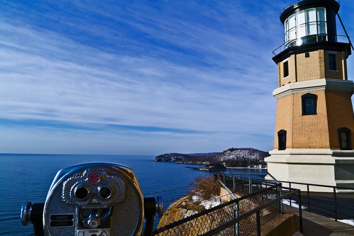 Split Rock Lighthouse on Lake Superior. Dramatic clouds stream past the light house with a calm lake and jagged cliffs in the background. In the foreground is a coin operated binocular.