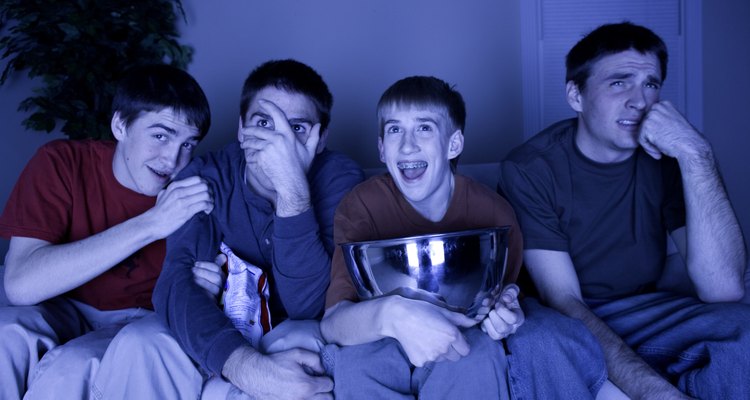 Four teenage boys (13-14) (16-17) sitting on couch watching TV, eating snacks