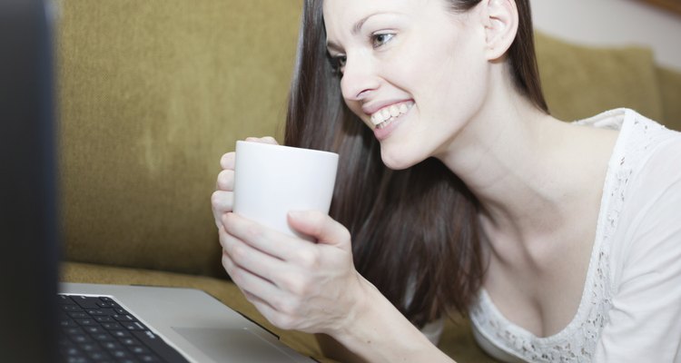 A woman holding a hot cup of tea while smiling at her computer screen.