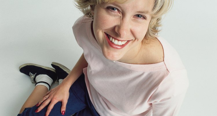 beautiful young slender blonde caucasian adult female wearing jeans and a  pale pink t-shirt and tennis shoes sits on the floor as she leans forward smiling at the camera playfully