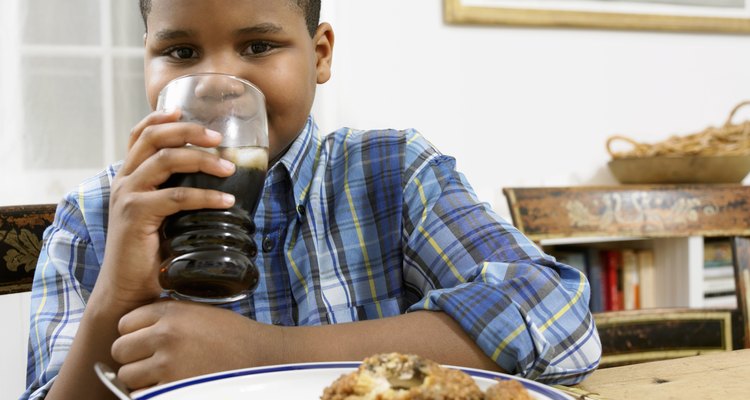 Young Boy Sits at a Table With a Plate of Food, Drinking a Glass of Cola