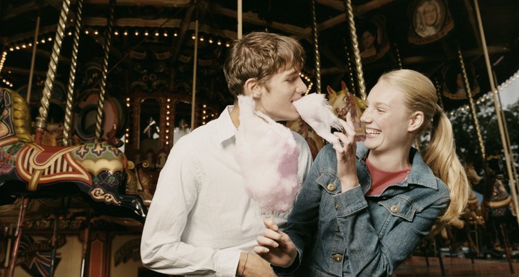 Teenage Couple Stand in Front of a Carousel, Girl Feeding Her Boyfriend Candy Floss and Laughing