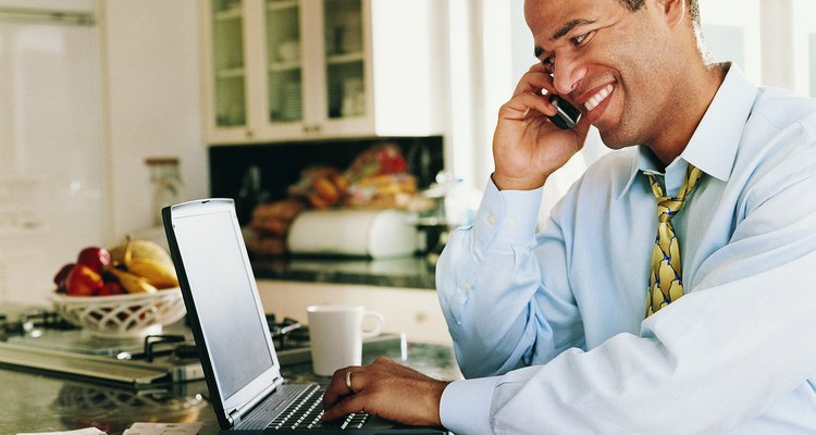 Man Sitting in a Kitchen Typing into a Laptop and Listening to his Mobile Phone