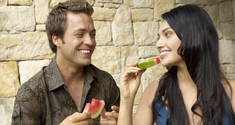Couple eating melon slices outdoors, looking at each other, laughing