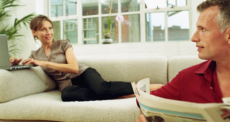 Man holding newspaper looking at woman on sofa using laptop