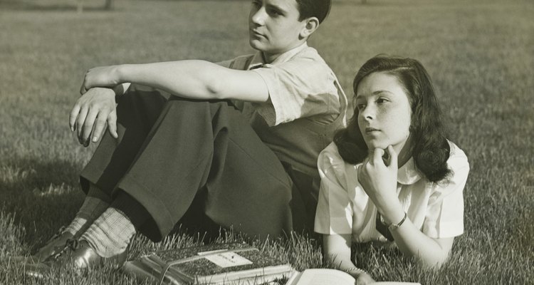 Two teenagers resting in field, boy (16-17) sitting, girl (14-15) lying with book, (B&W)