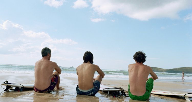 Three men sitting on wet sand by surfboards, rear view