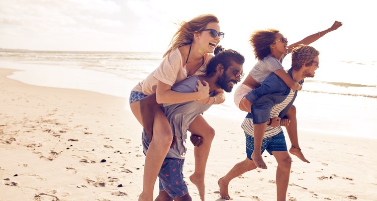 Two young men giving their girlfriends piggyback rides at the beach. Cheerful young friends enjoying summertime on the beach.