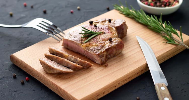 Tasty homemade well-done steak on wooden cutting board with fork and knife on stone background.