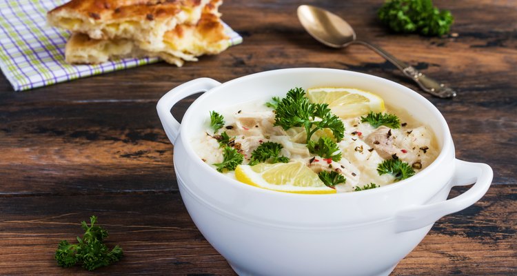 Avgolemono, chicken soup with egg-lemon sauce, rice and fresh parsley leaves in white bowl and freshly baked bread on wooden table.