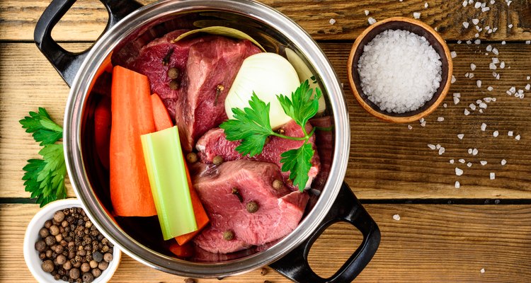 Ingredients for meat broth in pan on wooden table: beef, onion, carrot, celery, parsley and spices.