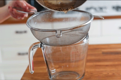 A cook pouring glaze through a fine-mesh sieve and into a glass measuring cup.