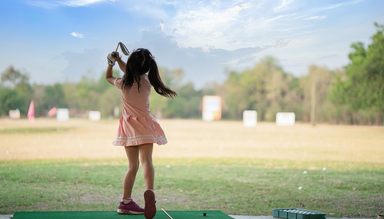 young girl practices her golf swing on driving range, view from behind