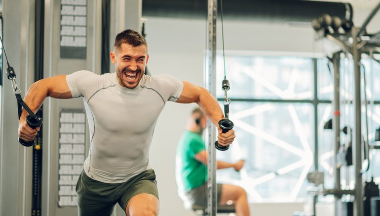 A sportsman is shouting in a gym while lifting heavy weights on a cable machine in a gym.