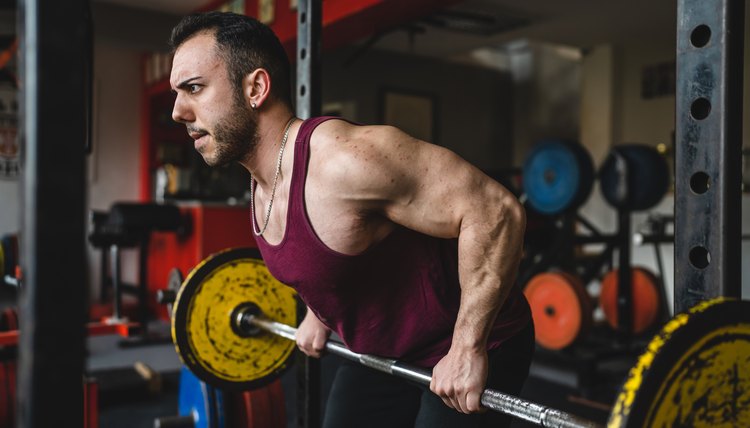 one man side view of adult male bodybuilder standing at the gym lifting weights with barbell and plates exercise back muscles doing rows for latissimus dorsi muscles