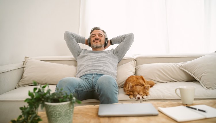 Calm Middle age Caucasian man sitting on sofa listening to music enjoying meditation for sleep and peaceful mind in wireless headphones, leaning back with his lovely chihuahua dog sit besides.