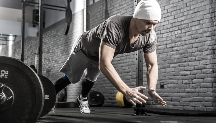 Strong guy with a beard makes a pushup with a front clap in the gym on the gray brick wall background. He wears sportswear, white sneakers and a white cap. Man looks in front of himself. Horizontal.
