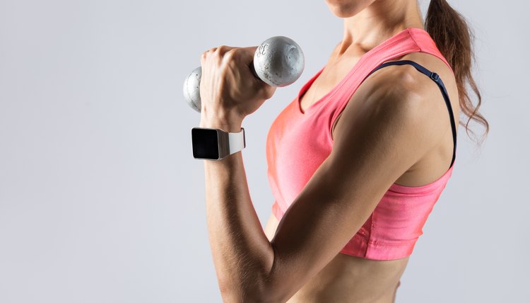 Close-up of torso of beautiful young fitness person wearing smartwatch and red sportswear top holding dumbbell. Sporty model girl working out, doing weight training with dumbbells on grey background