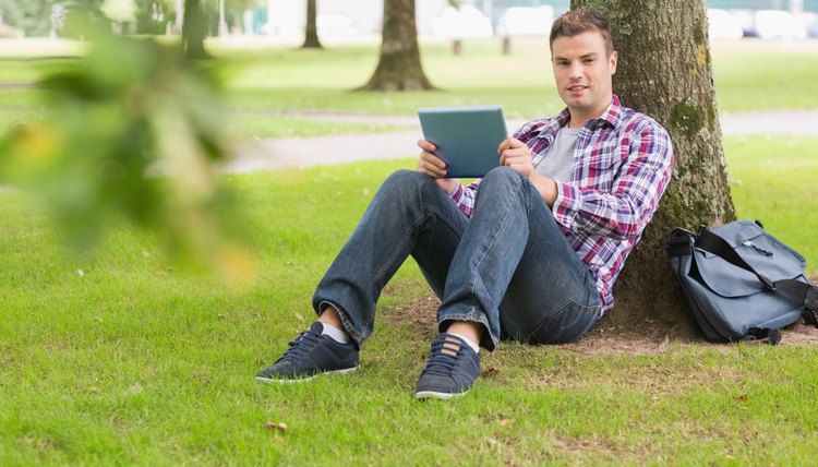 A leather messenger bag beside man leaning against tree.