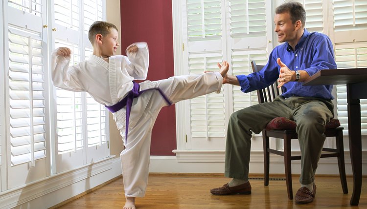 A boy in a martial arts uniform practices a side kick into his father's hand