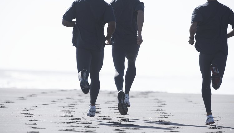 Three men jogging on beach, low section, rear view