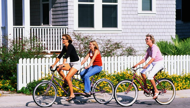 adult woman and teenage girl riding a bicycle built for two down a street by a beach house, mature adult female is behind them also riding a bike. 