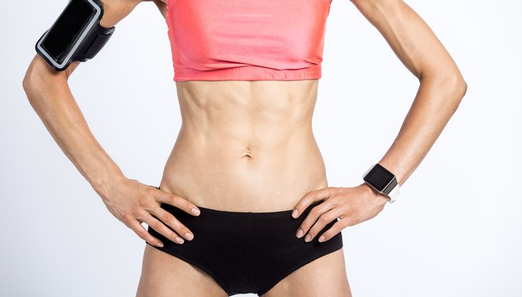 Closeup of torso of beautiful young person wearing armband with smartphone, smartwatch, shorts and red sportswear top. Sporty model girl with perfect abs, slim waist and hips working out. Studio image