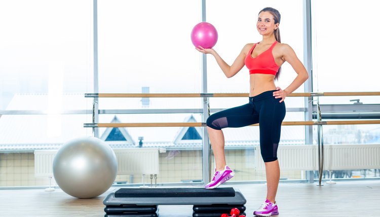 Fitness girl, wearing in sneakers, red top and black  breeches, posing on step board with ball, on the sport equipment background, in the gym.