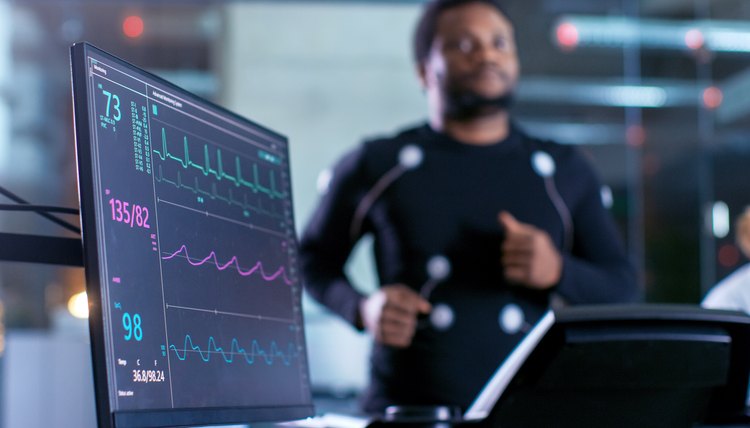 Close-up Shot of a Monitor With EKG Data. Male Athlete Runs on a Treadmill with Electrodes Attached to His Body while Sport Scientist Holds Tablet and Supervises EKG Status in the Background.