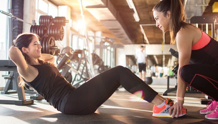 Young woman exercising sit-ups with assistance of female friend in gym.